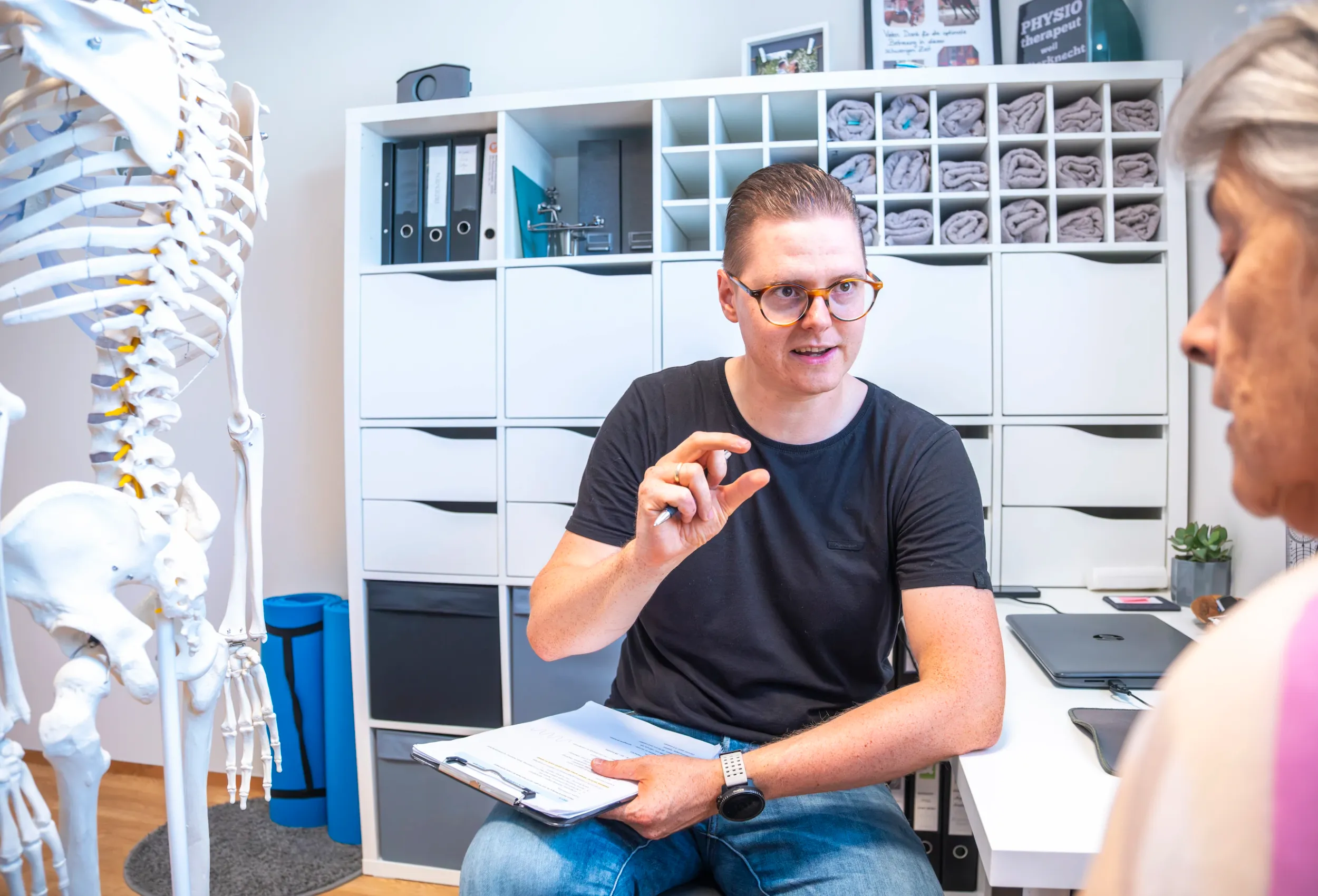 Therapist with glasses explaining something to a patient in the consultation room, with a skeleton model and a shelf of folders in the background.