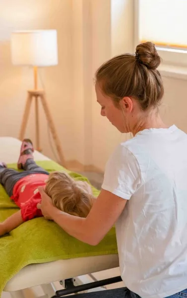 Woman with hair in a bun massaging child lying on a table covered with a green cloth in a bright room with a floor lamp