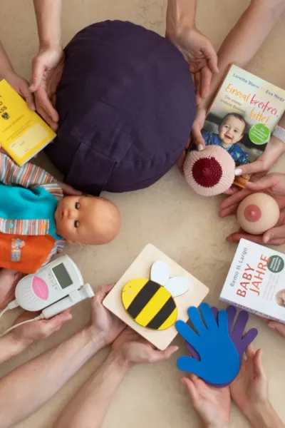 Top-down view of a circle of hands holding various pregnancy and family related items in the centre, including a baby doll, a breastfeeding model and reference books.
