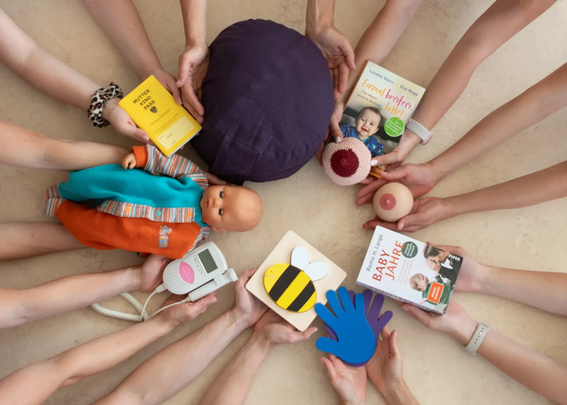 Many hands reach from outside into a circle holding items such as a mother-child passport, books, a breastfeeding model, a CTG device and toys, viewed from above.