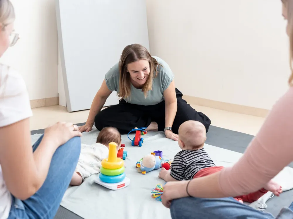 Course instructor sitting smiling among mothers and babies playing with toys on a blanket