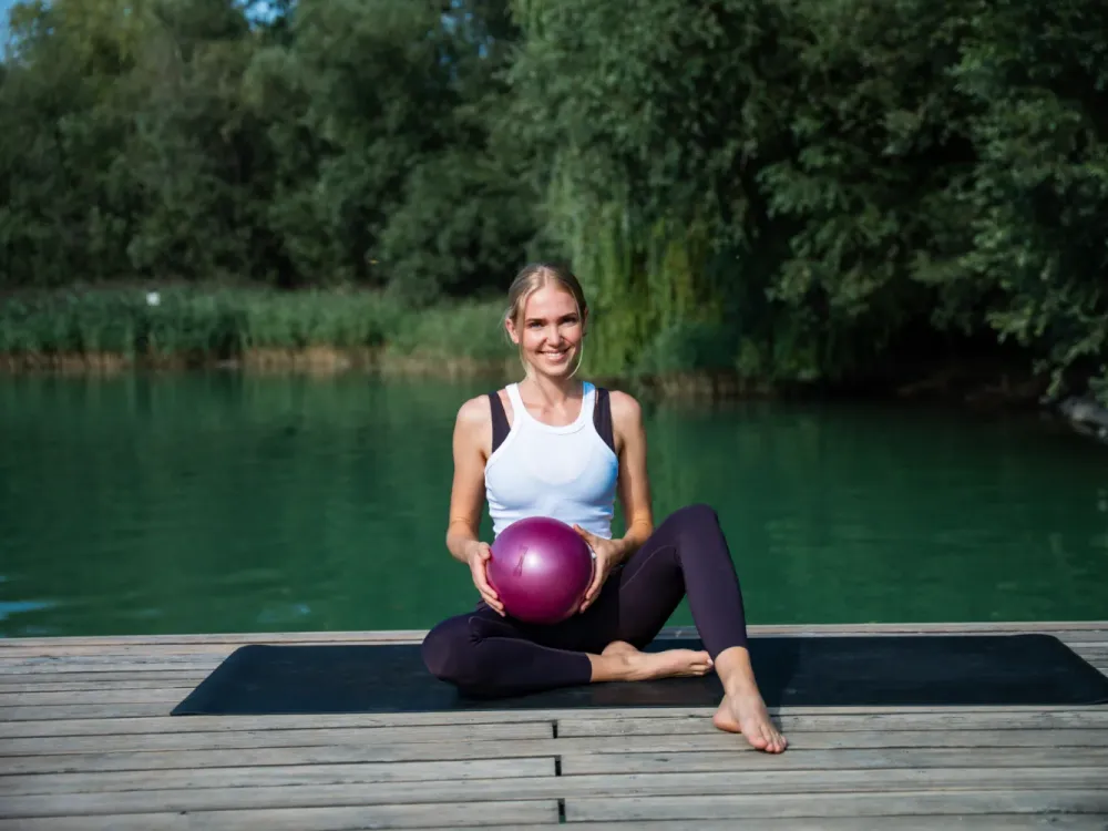 Woman sitting smiling on a jetty by a lake holding a pilates ball