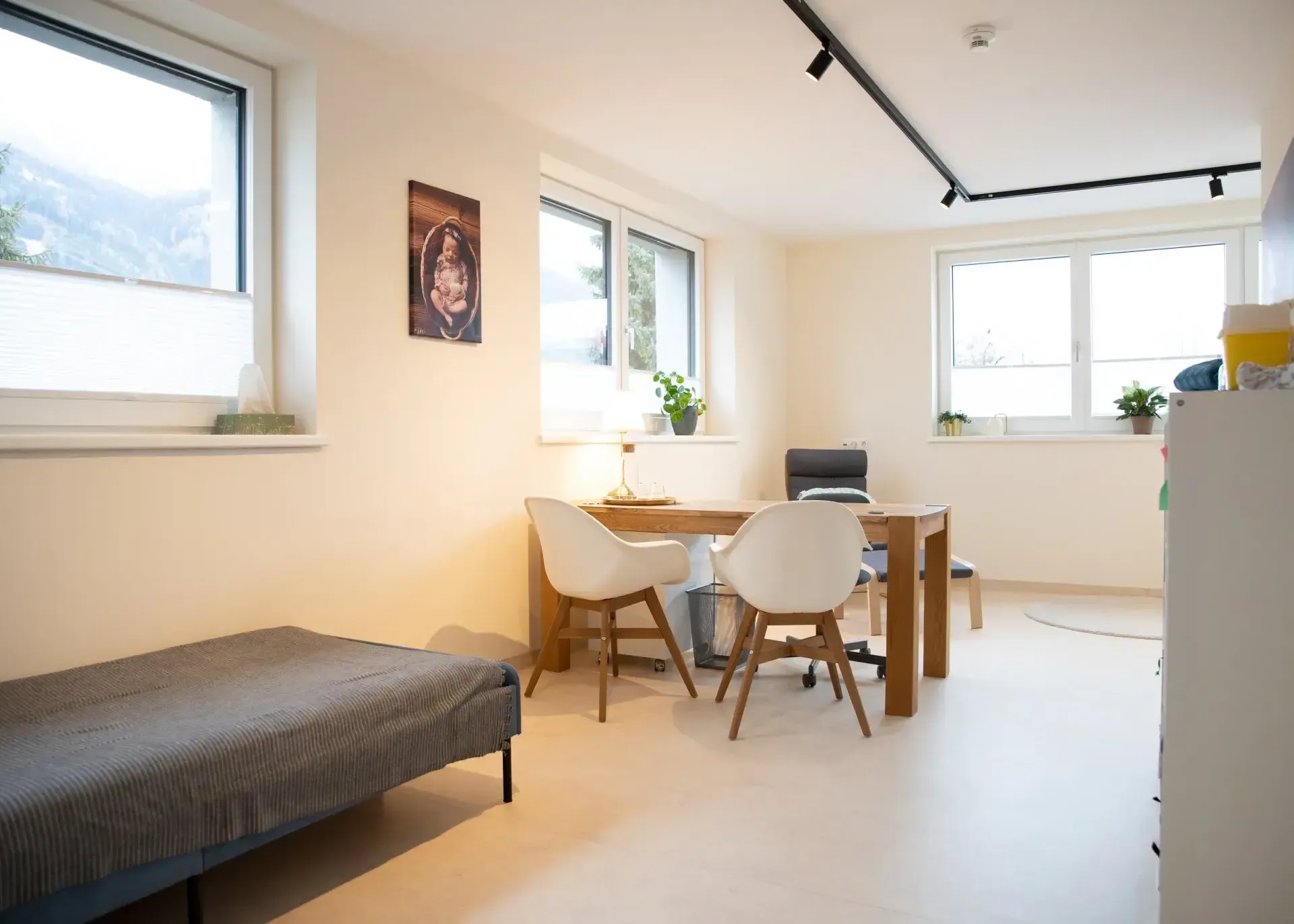 Heller Raum mit Esstisch, zwei Stühlen, einem Bett und großen Fenstern mit Blick auf Berge.