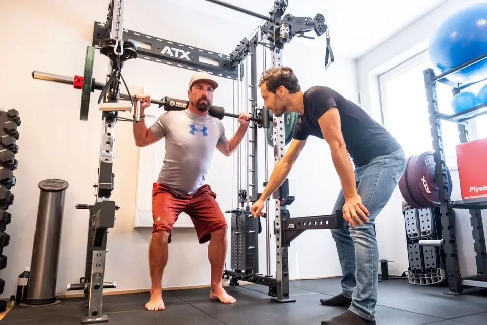 Trainer unterstützt einen Mann beim Kniebeugen-Training mit Langhantel im Kraftraum mit ATX-Trainingsgeräten.