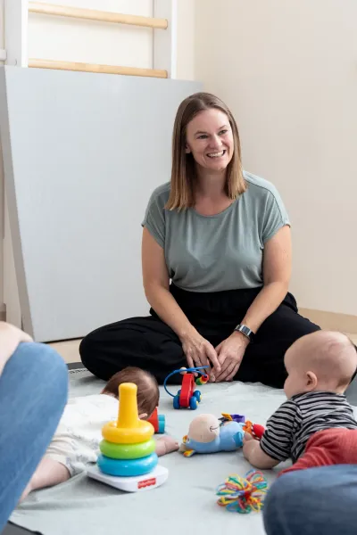 Course instructor sitting cross-legged and smiling on a mat between two mothers, while two babies play with toys on a blanket